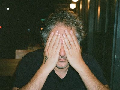 Harmony Korine has his eyes covered by his hands and his trident tattoo is visible on his right hand.